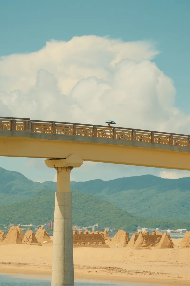 Pedestrian bridge framing sand sculptures with mountains and clouds beyond
