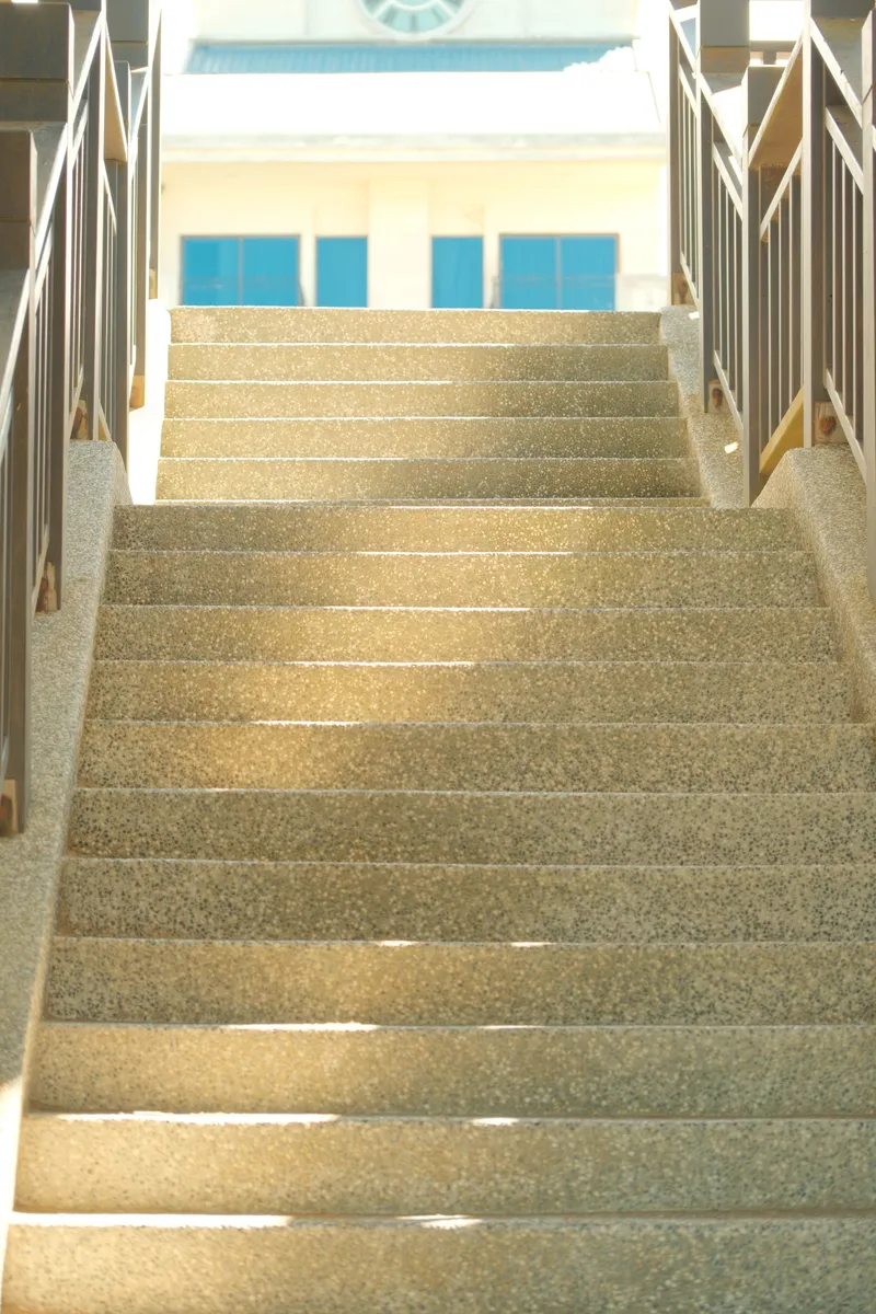 Sunlit granite stairway flanked by metal railings leading toward a building