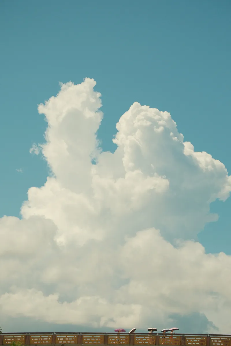 Towering cumulus clouds rising above a bridge with tiny umbrellas visible below